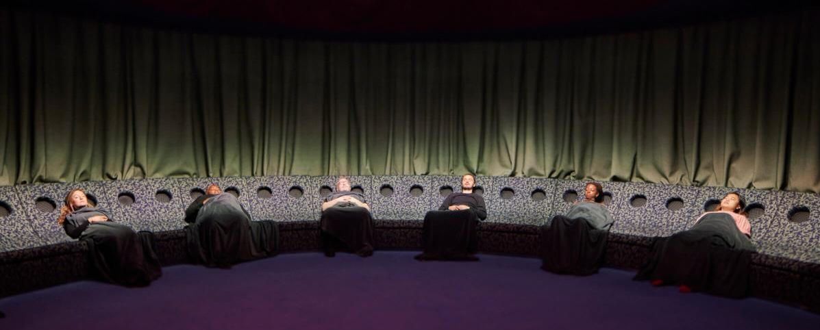 Six people recline with their eyes closed around the edge of a circular Dreamachine installation, beneath a large glowing pink light. Six people recline with their eyes closed around the edge of a circular Dreamachine installation, beneath a large glowing pink light.