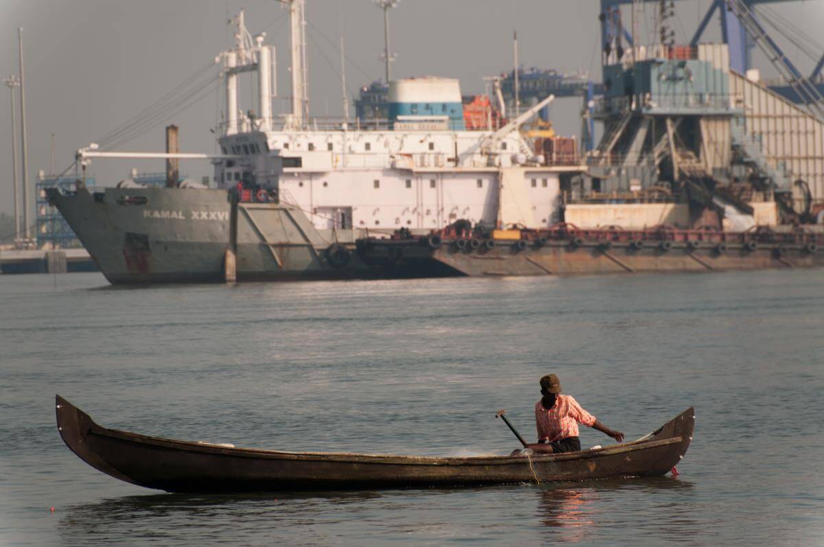 A person in a small wooden fishing boat on calm water, with a large industrial ship and port infrastructure in the background.