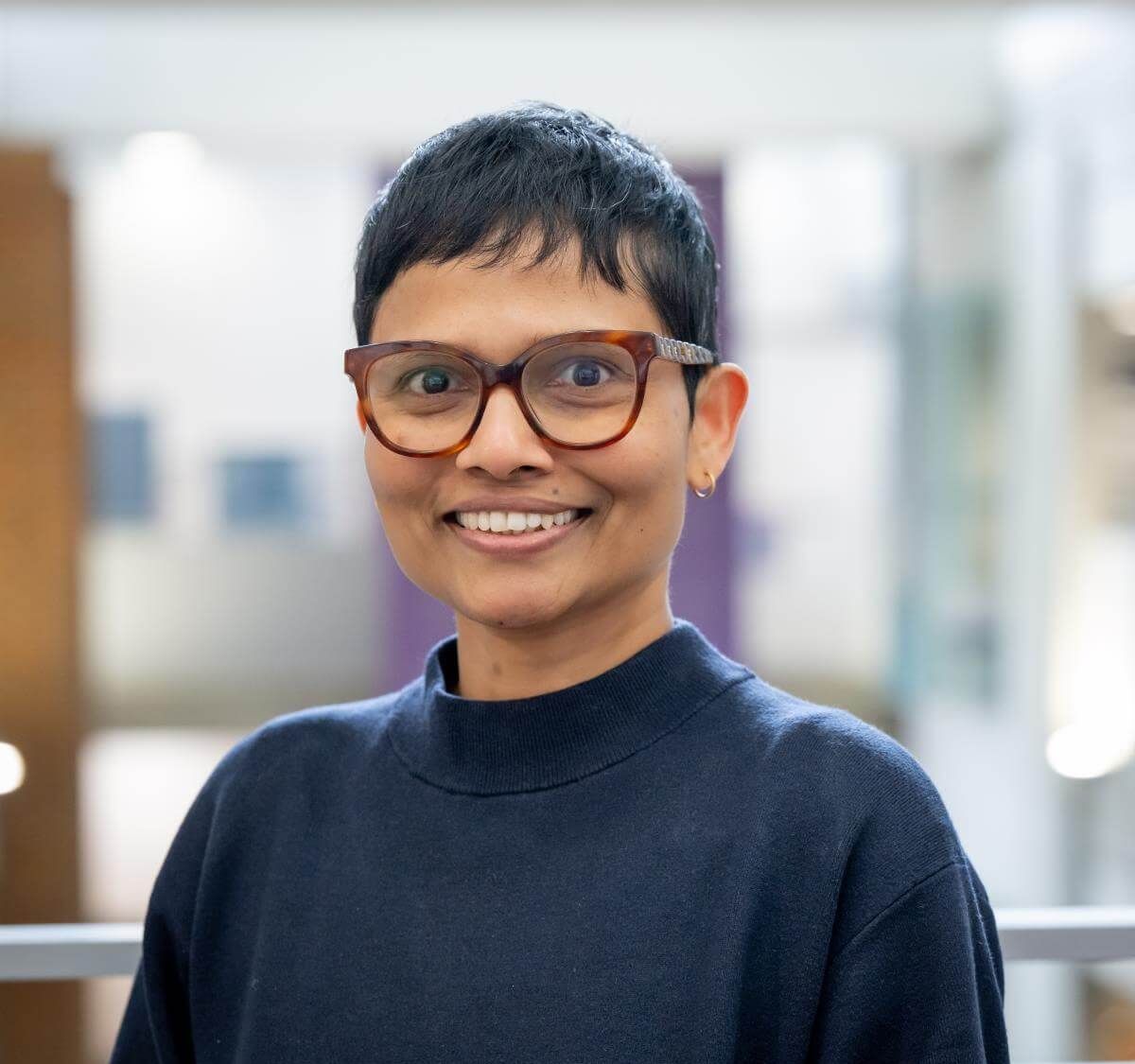 Dr Shyamain Wickramasingha smiling at the camera, wearing glasses and a navy jumper, against a softly blurred indoor background.