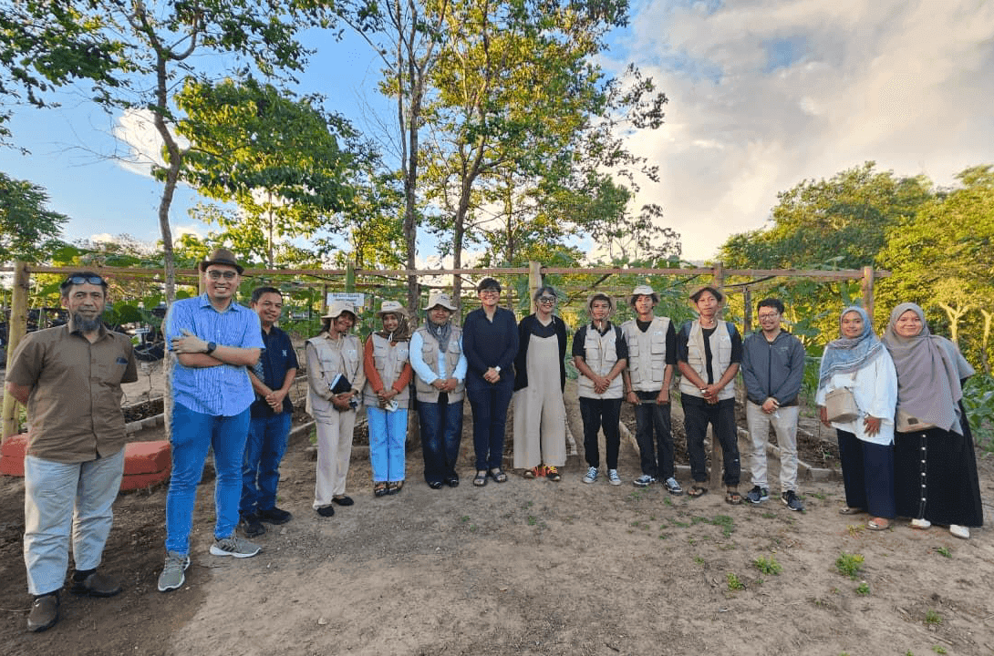 Fourteen researchers stand in a line for a group photo on a farm plot, some wearing field vests and sun hats, with trellised crop rows, trees and a bright sky in the background.