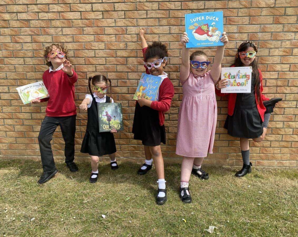 Five children in school uniform pose in front of a brick wall wearing colourful superhero-style glasses and holding up books, with one child raising a book above their head.