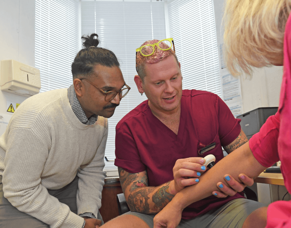 Two clinicians in a surgery examine a patient’s forearm; one, in medical scrubs, uses a handheld device on the patient’s skin while the other observes closely, illustrating collaborative healthcare and clinical research.