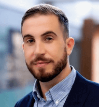 Head shot of Rocco d’Este, Associate Professor in Economics, wearing a blue blazer and checked shirt, looking at the camera with a neutral expression.