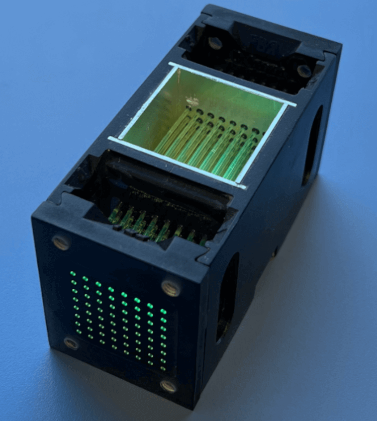 Close-up of the LiquidO detector: a black rectangular module with a small window revealing bundled optical fibres inside, and a grid of green indicator lights on the front.