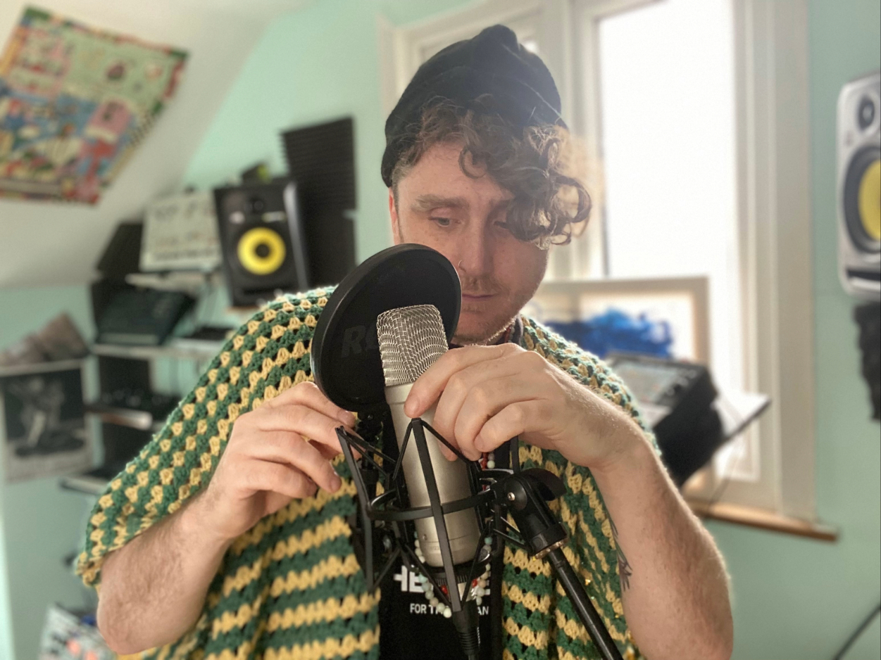 Ben Kelly stands in a light-filled home studio, wearing a knitted top whilst adjusting a condenser microphone and pop filter. 