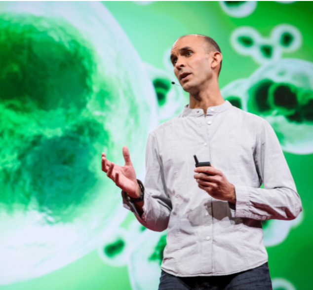 Professor Anil Seth on stage delivering a lecture. He gestures with one hand and holds a presentation remote in the other; behind him, a vivid green slide shows enlarged, cell-like shapes.