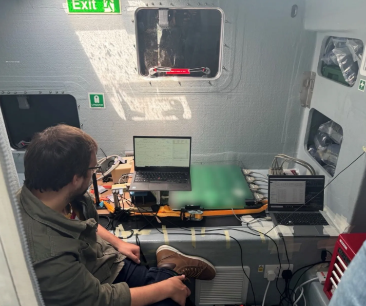 Tom Coussens sits in a ship’s cabin monitoring two laptops wired to a green quantum magnetometer test rig during Royal Navy sea trials.