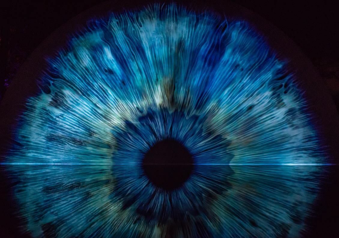 Large-scale projected artwork of a blue human iris, with radiating fibres around a dark pupil, shown against a dark background.