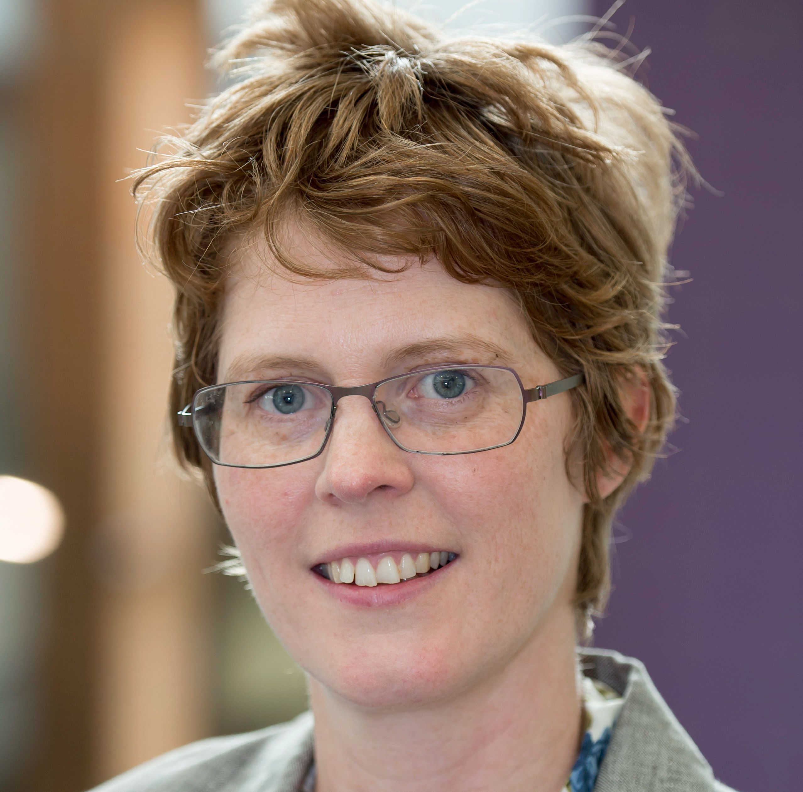Professor Annemie Maertens, smiling, wearing glasses and a grey jacket over a patterned blouse.