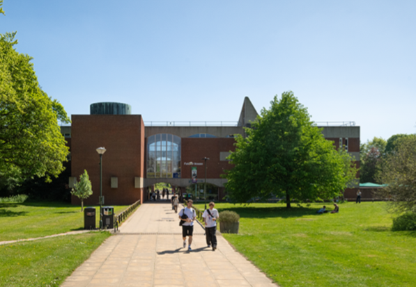 Students walking along a wide path on the University of Sussex campus, heading toward Falmer House on a sunny day, with trees and green space on either side.