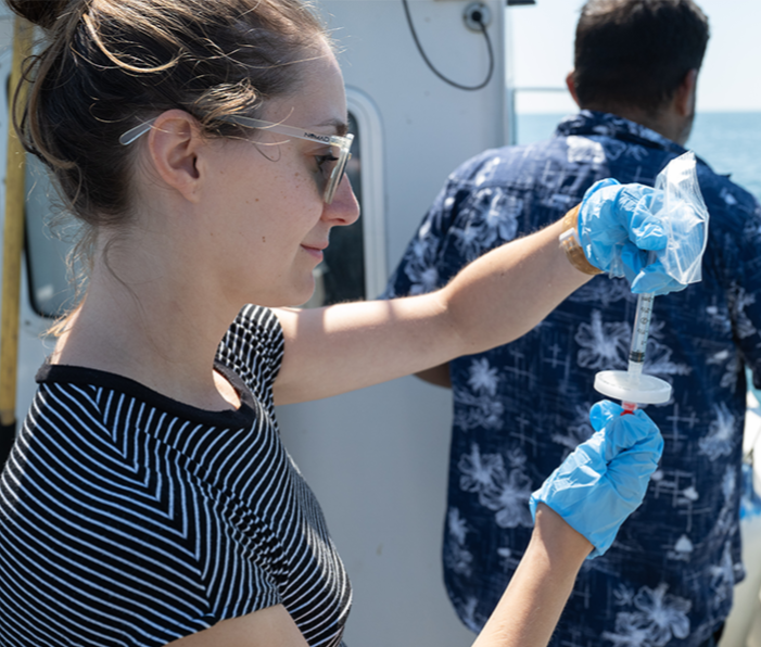 Alice Clark collects an environmental DNA (eDNA) sample from seawater while on a boat, wearing gloves and sunglasses.