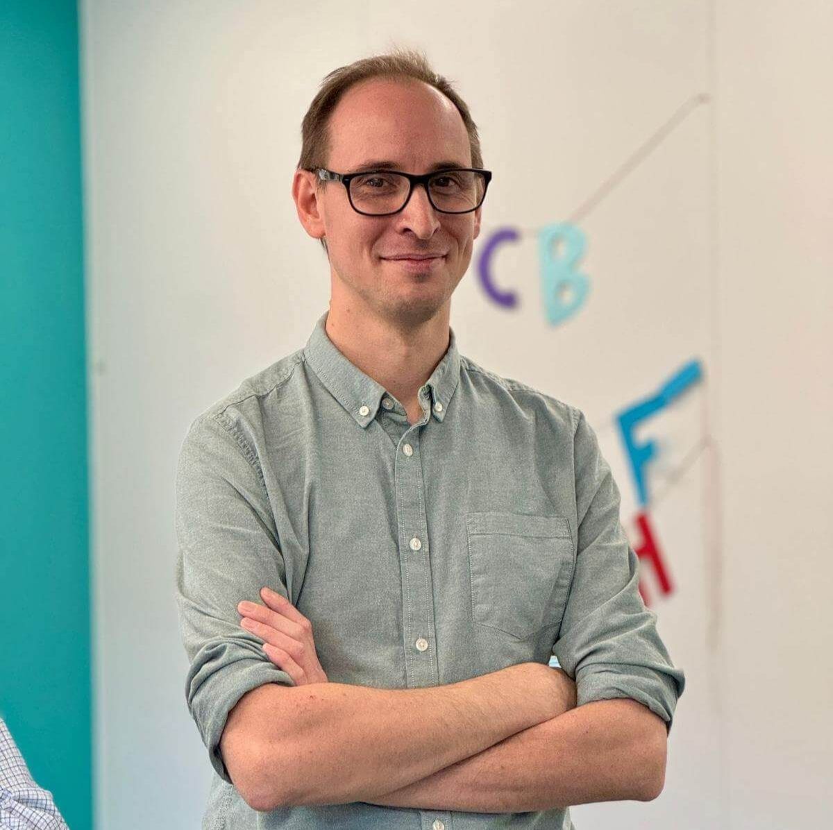 Dr Liam Berriman stands indoors with arms crossed, wearing glasses and a light green button-up shirt, with colourful letters blurred on the wall behind him.