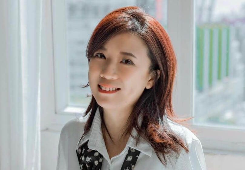 Karrie Liu smiles at the camera indoors by a bright window, wearing a white blouse with a patterned scarf.