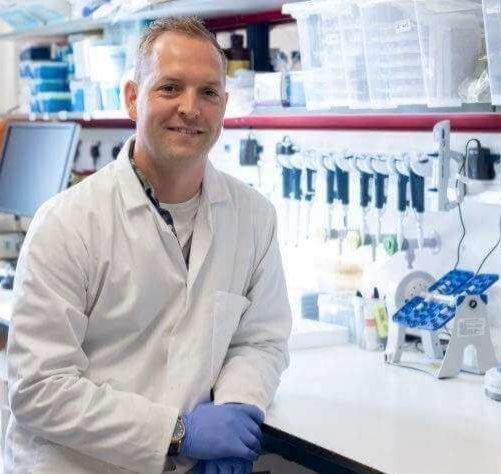 Dr Rhys Morgan sits at a laboratory bench wearing a white lab coat and blue gloves, with lab equipment in the background.