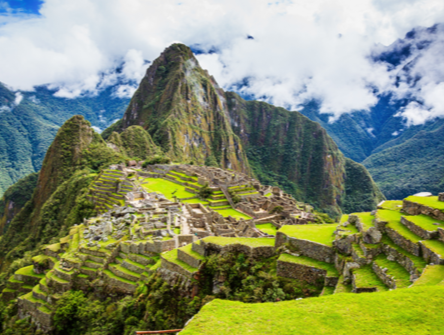 Panoramic view of Machu Picchu, the ancient Inca citadel in the Andes Mountains of Peru, with stone terraces and ruins set against steep green peaks and a cloudy sky.