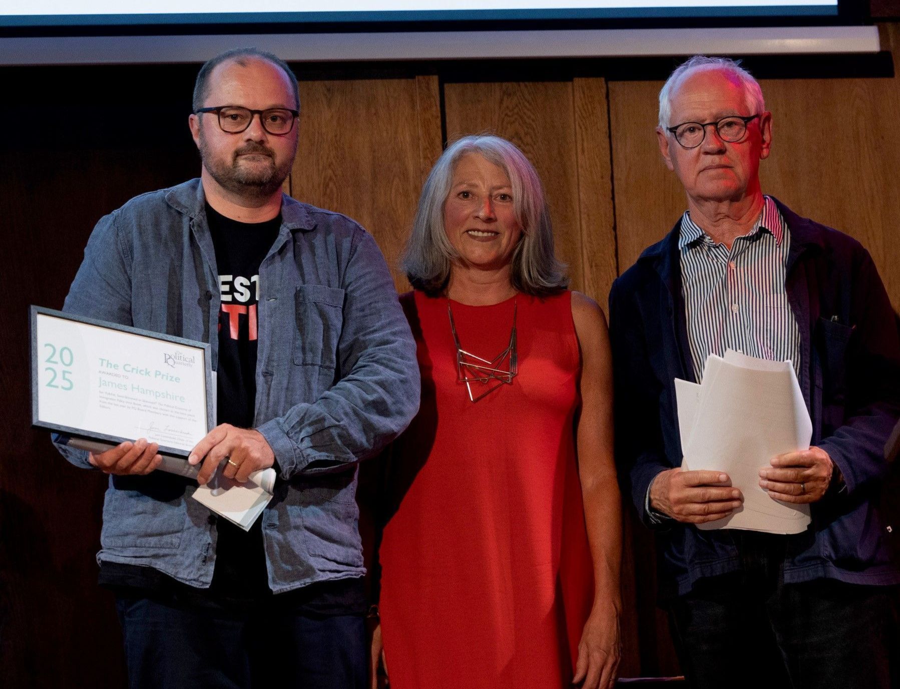 Professor James Hampshire (left) holding his framed 2025 Bernard Crick Prize certificate, standing with two other individuals on stage at the Orwell Prize ceremony.