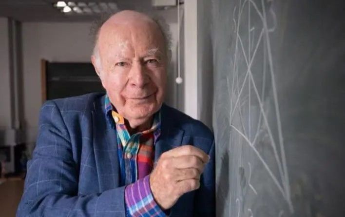 Professor James Hirschfeld standing at a chalkboard, holding chalk, with geometric diagrams visible. He is wearing a colourful shirt and blue blazer, looking warmly at the camera.