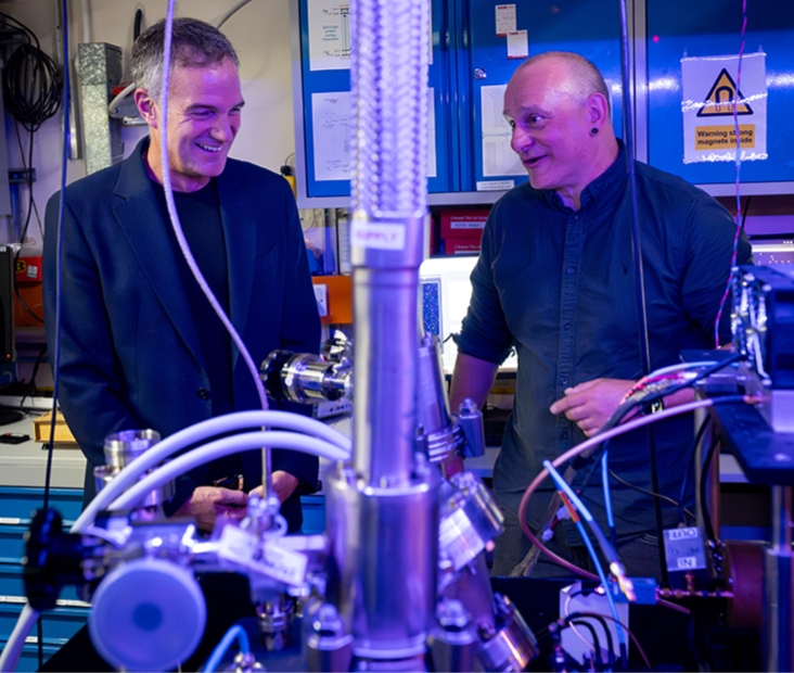 Peter Kyle, Secretary of State for Science, Technology and Innovation (left), and Professor Winfried Hensinger (right) talking together in a quantum technologies lab at the University of Sussex, with research equipment in the foreground.