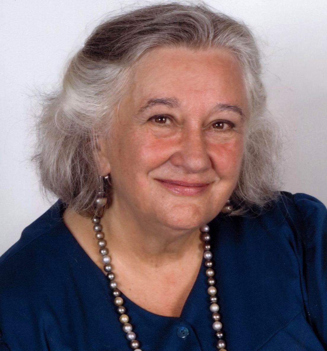 Professor Margaret (Maggie) Boden smiling warmly at the camera, wearing a navy blue blouse and beaded necklace.