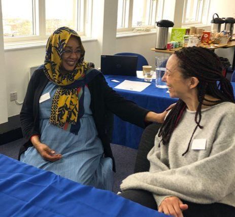 Two women seated at a table, smiling and talking during a community or workshop event. One wears a patterned headscarf and blue dress, the other a grey jumper with long braids.