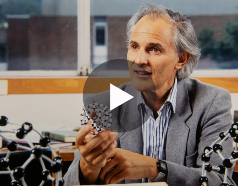 Video thumbnail showing scientist Sir Harry Kroto in an office holding a ball-and-stick model of C60, with a large play icon overlaid.