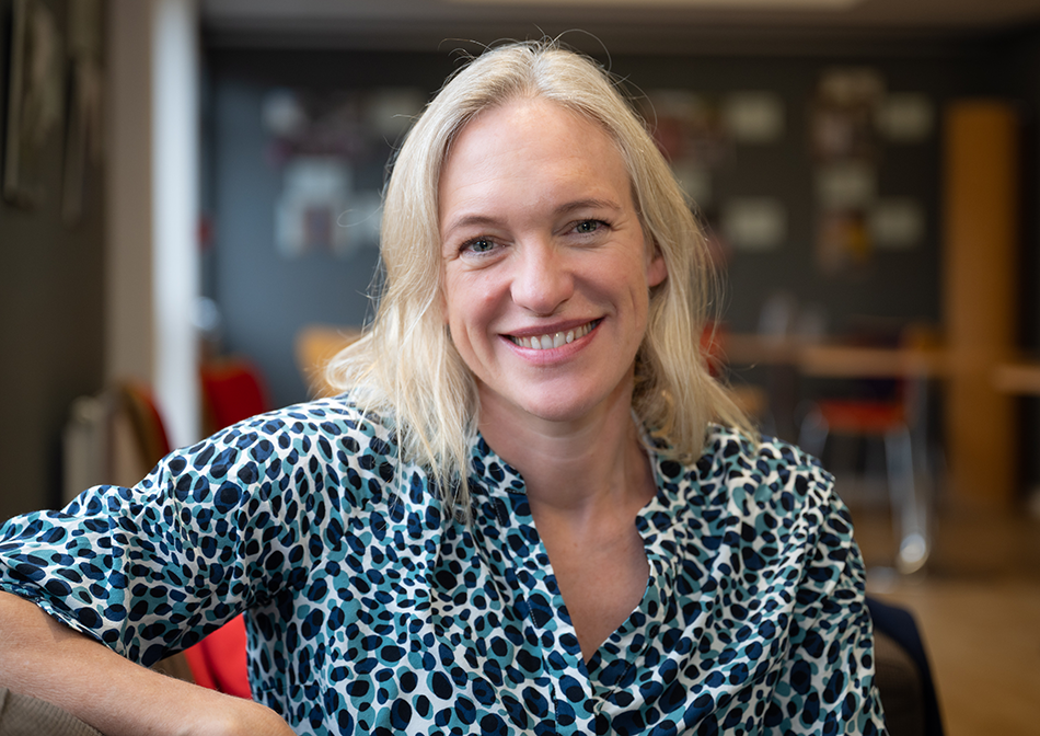 Dr Sarah Stephens smiling at the camera, seated indoors in a softly lit room. She has shoulder-length blonde hair and is wearing a black and white patterned blouse.