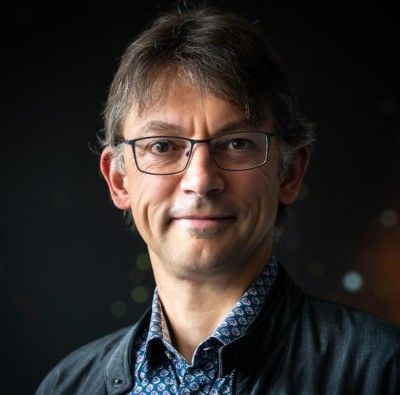 Professor Seb Oliver, smiling, wearing glasses, a patterned shirt, and a dark jacket.
