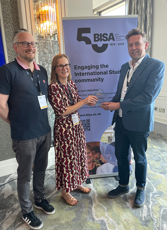 Peter Newell (right) receiving his award at the 2025 BISA conference in Belfast. He stands smiling with two other people in front of a BISA 50th anniversary banner that reads “Engaging the International Studies community.”