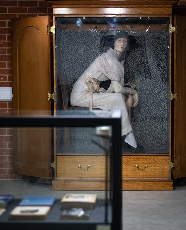 Lifelike figure of Virginia Woolf seated in an open wooden wardrobe, with books displayed in a nearby glass case. Part of the Pressing Matters exhibition at the University of Sussex Library.