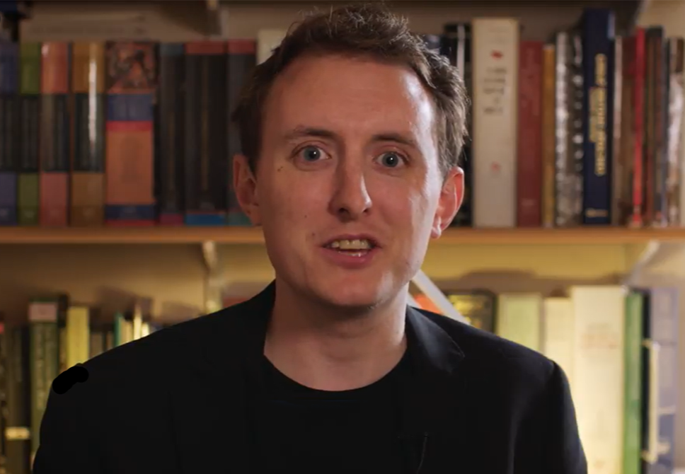 Tom F Wright speaking to camera in front of a book-filled background, wearing a dark jacket.